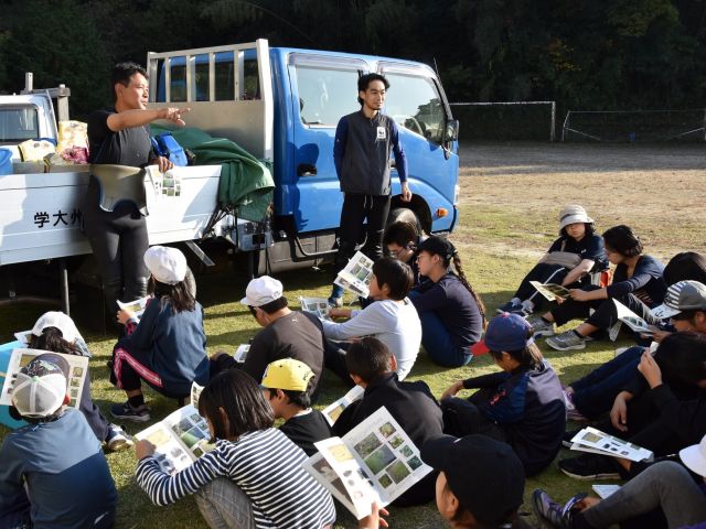 水田・水路の生物多様性と農業の共生プロジェクトの一環で、プロジェクトサイト熊本県玉名市において、地元の子ども会とおこなった生きもの観察会の様子。共同研究者の大学准教授から淡水魚の説明を受ける子供たち。